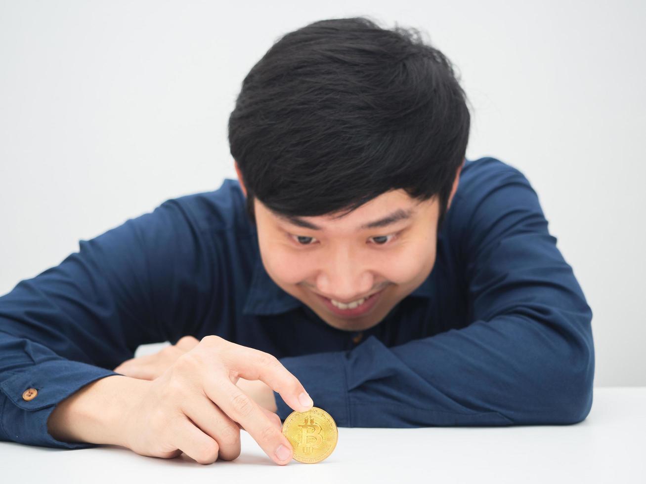 asiatischer mann, der am schreibtisch sitzt und goldene bitcoin in der hand mit einem lächeln betrachtet foto