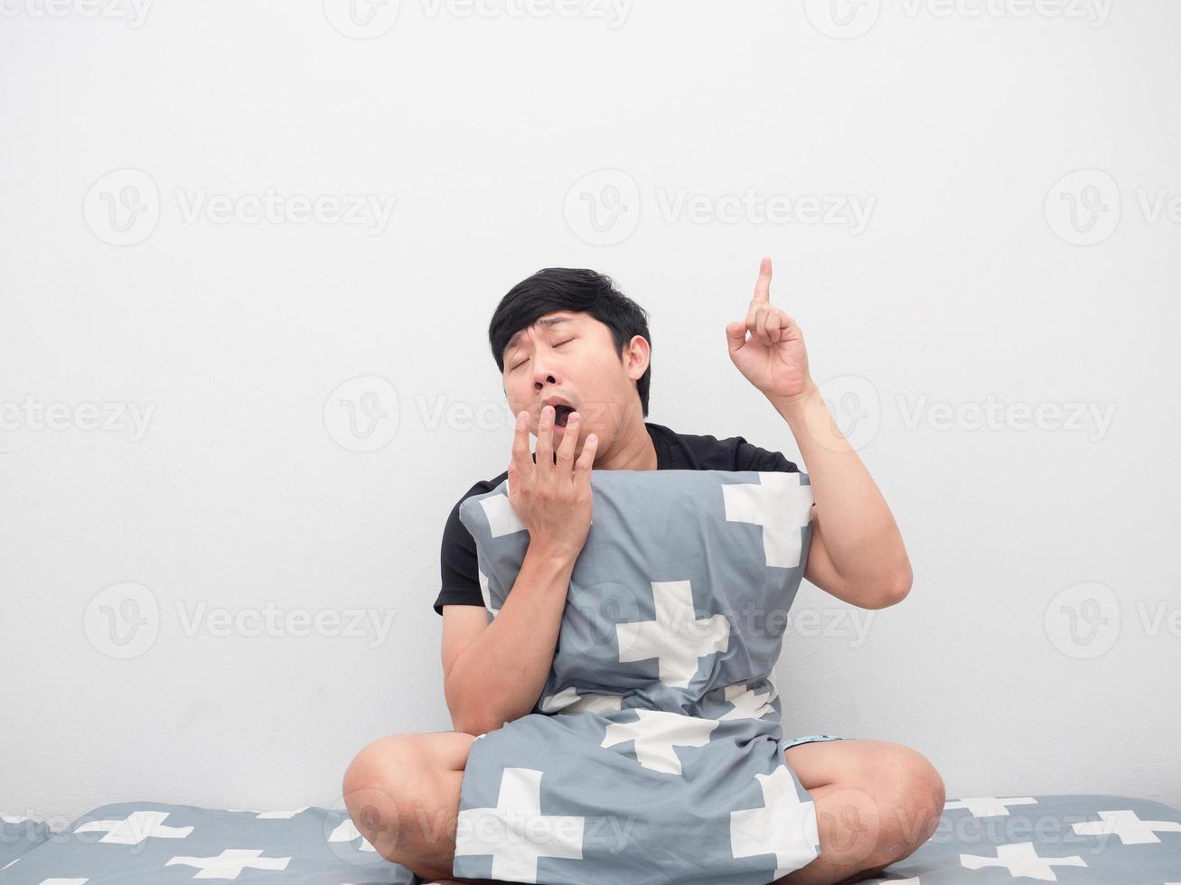 Schläfriger Mann mit Kissen auf dem Bett gähnt und zeigt mit dem Finger nach oben foto