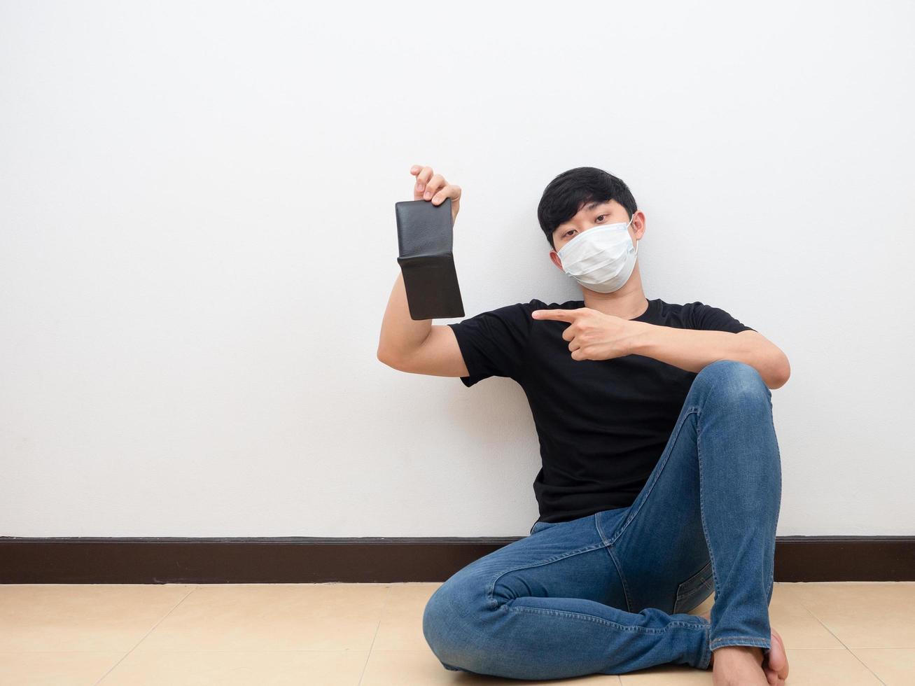 Der arme Mann mit Maske zeigt mit dem Finger auf eine leere Brieftasche, die auf dem Boden sitzt foto