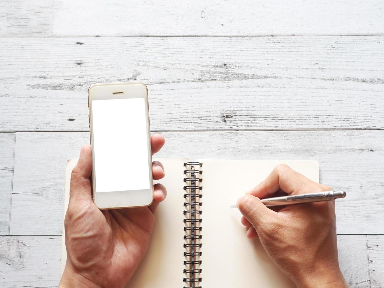 Hand, die das weiße Display des Smartphones hält, und mit einem silbernen Stift auf einem weißen Holztisch mit Blick auf den Naturschatten auf das Notizbuch der leeren Seite schreiben foto