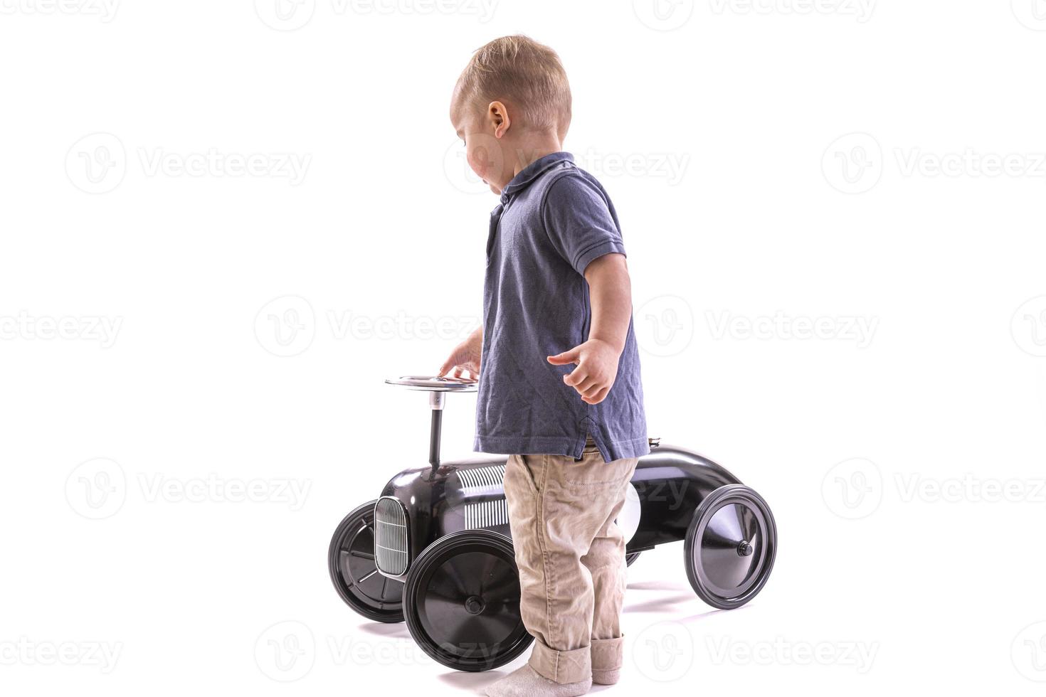 kleiner Junge, der Spielzeugauto im Retro-Stil fährt. Junge reitet altes Tretauto aus Metall für Kinder aus dem 19. Jahrhundert. isoliert auf weißem Hintergrund foto