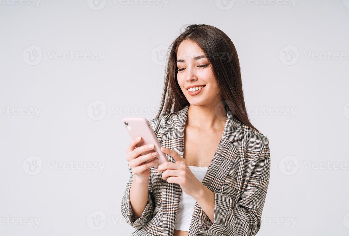Junge glückliche asiatische Geschäftsfrau mit langen Haaren im Anzug mit Handy auf grauem Hintergrund foto