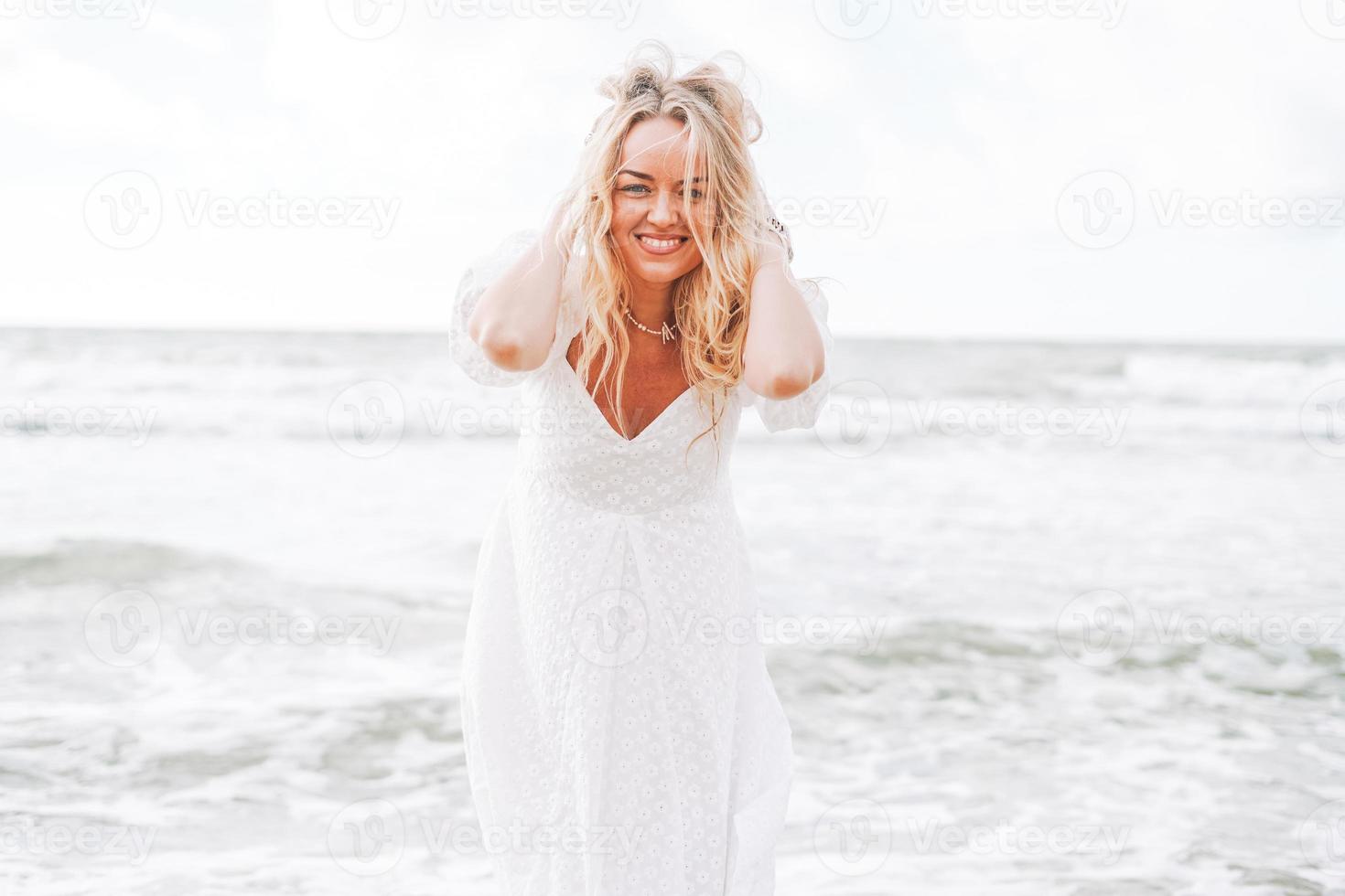 junge blonde schöne Frau mit langen Haaren in weißem Kleid, die das Leben am Meeresstrand genießt foto
