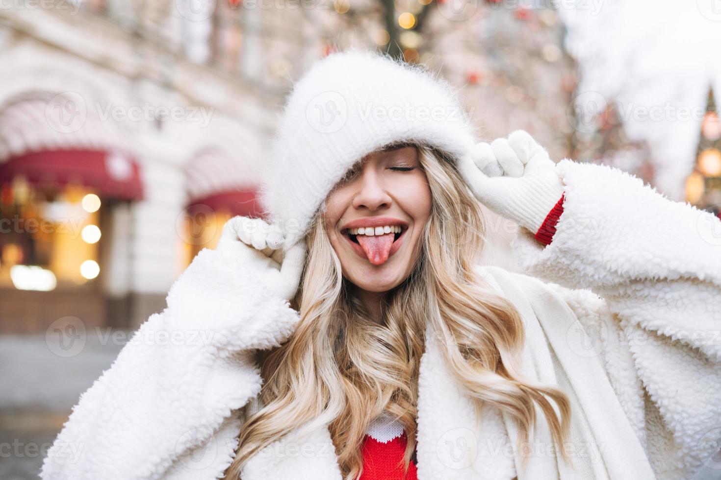 junge glückliche frau mit lockigem haar in weißer pelzmütze, die sich auf dem weihnachtsmarkt in der mit lichtern geschmückten winterstraße amüsiert foto