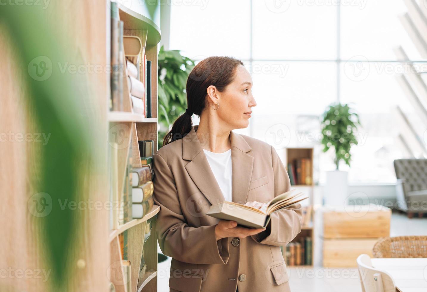 erwachsene lächelnde brünette geschäftsfrau vierzig jahre mit langen haaren in stylischem beige anzug und jeans an öffentlichem ort, grünes open space office, coworking. freundlicher lehrer oder mentor mit buch in der bibliothek foto