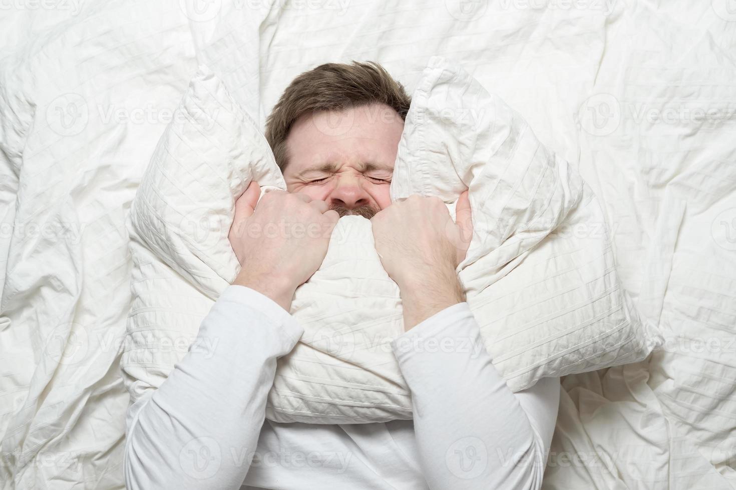 mann im weißen pyjama ist gestresst, hat schlaflosigkeit, kann nicht schlafen, bedeckt nervös den mund mit einem kissen und kneift die augen zu. Ansicht von oben. foto