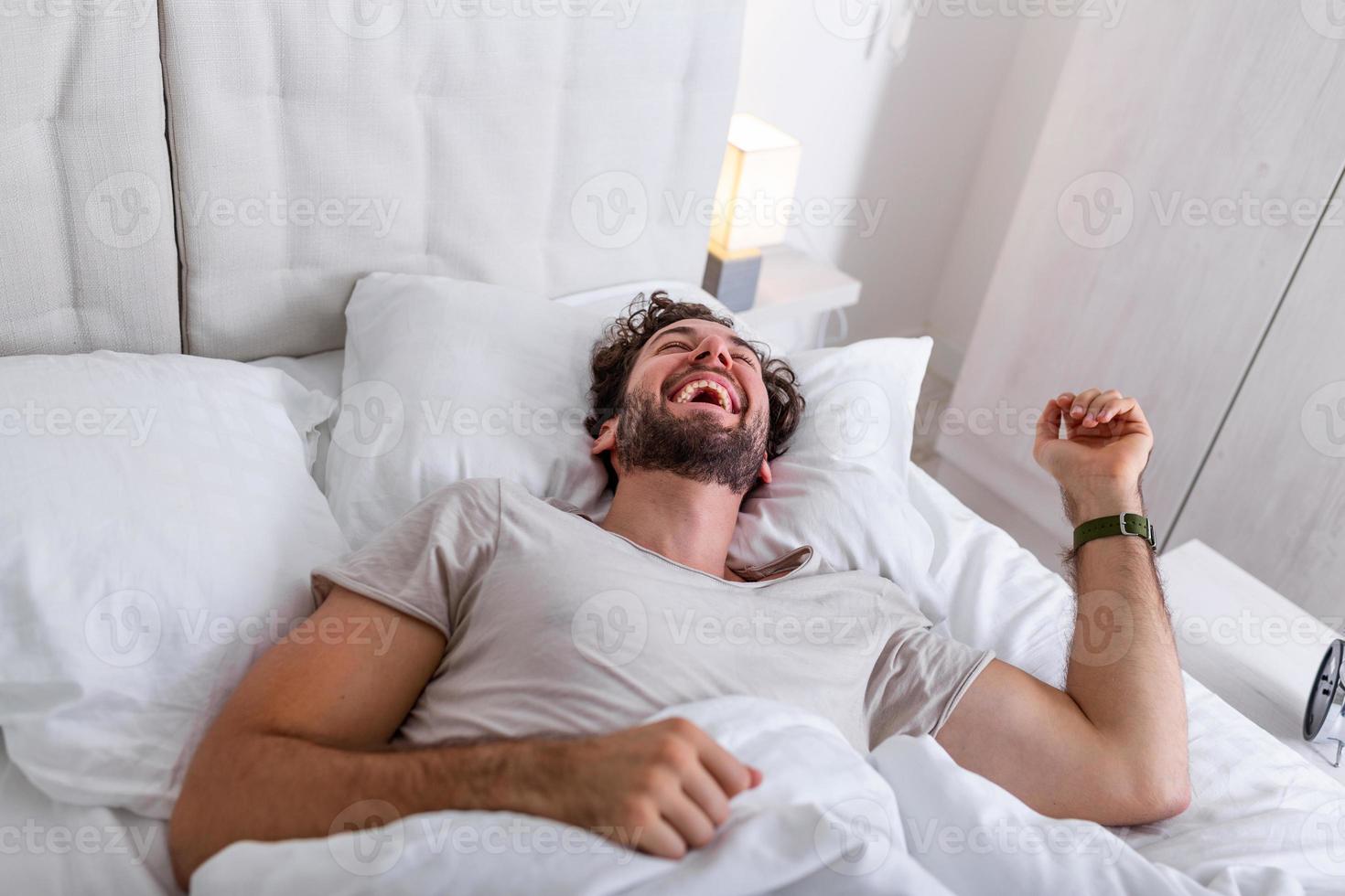 junger Mann schläft, wacht auf und streckt sich in seinem Bett. menschen, schlafenszeit und ruhekonzept - mann liegt zu hause im bett, es ist zeit aufzuwachen foto