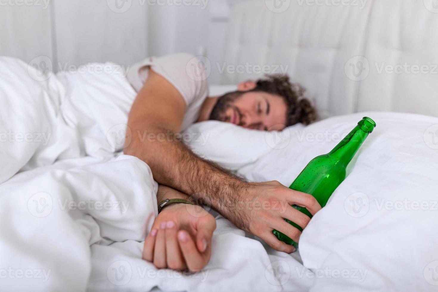 betrunkener mann im bett und trauriger ort und eine alkoholflasche in der hand. Junger Mann, der tödlich betrunken im Bett liegt und eine fast leere Flasche Schnaps hält. foto