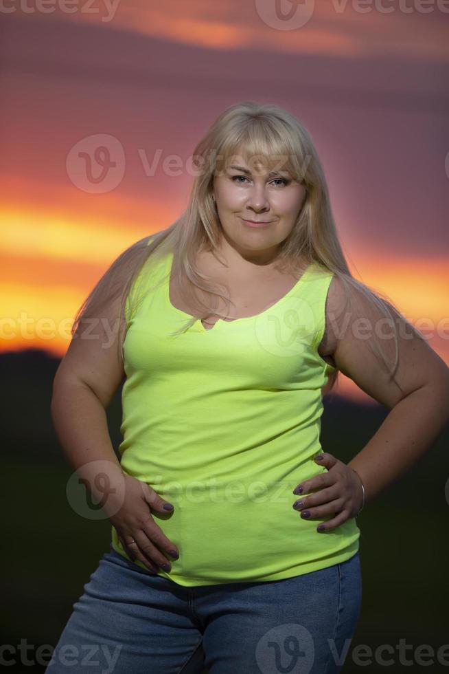 Eine mollige Frau mittleren Alters posiert in Jeans und einem hellen T-Shirt vor dem Hintergrund des Abendhimmels, übergewichtig xxl. ein volles Mädchen genießt das Leben. foto