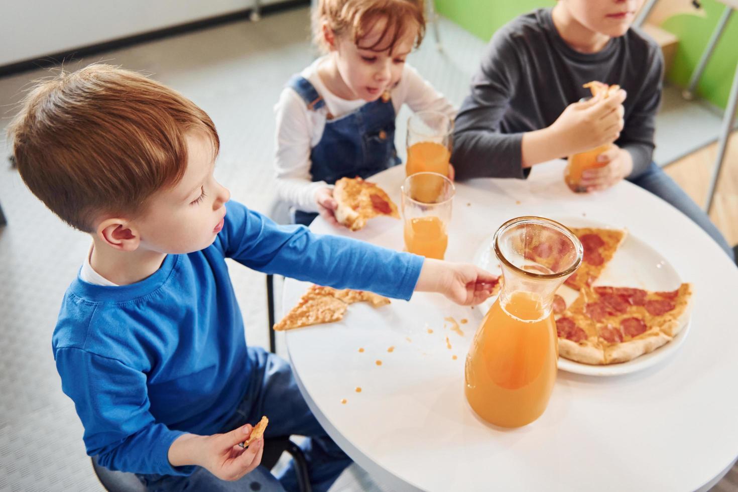 Drei Kinder sitzen drinnen am Tisch und essen gemeinsam Pizza mit Orangensaft foto