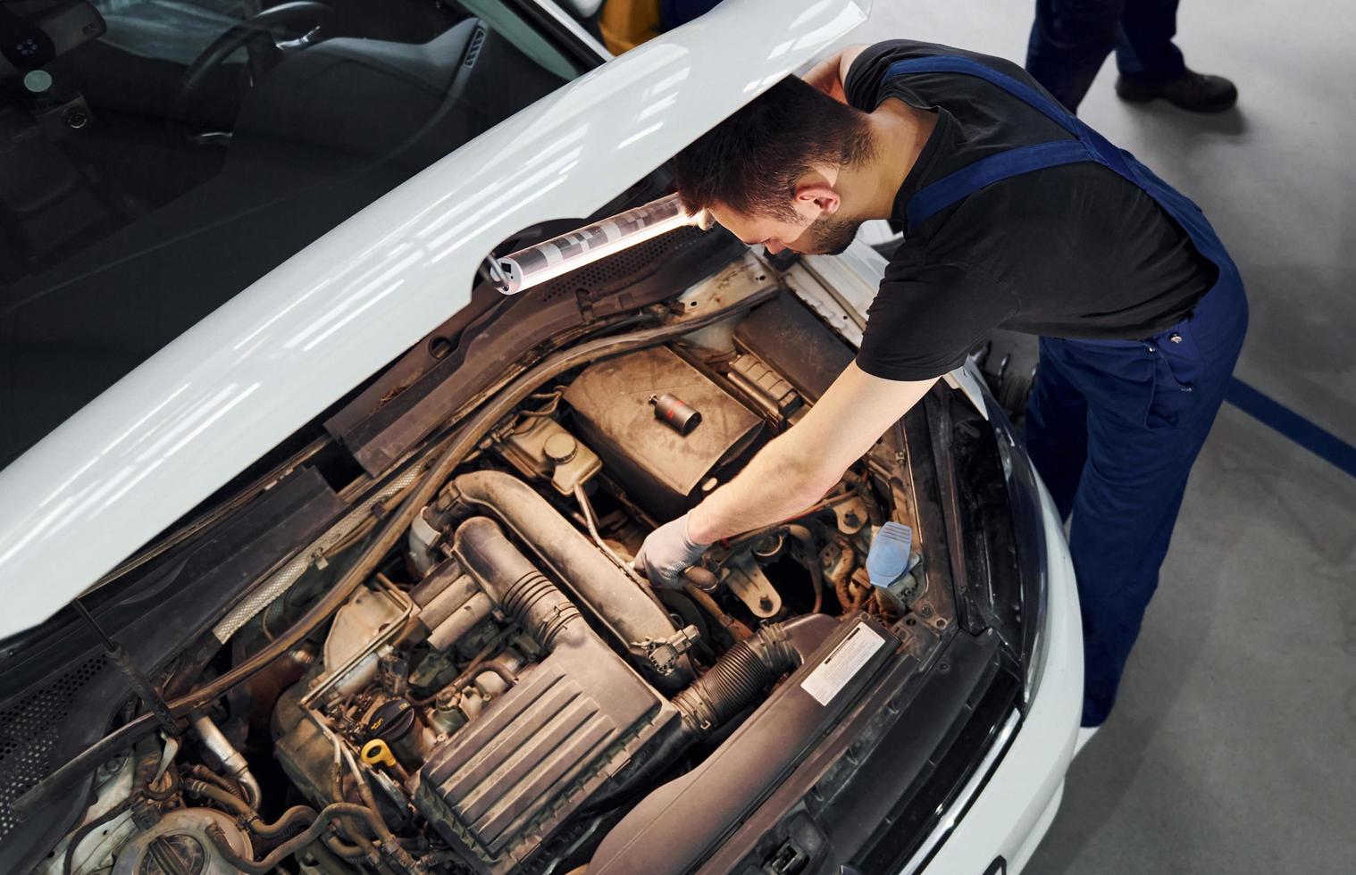 unter der Haube. Mann in Arbeitsuniform repariert weißes Auto drinnen. Konzept des Autoservices foto