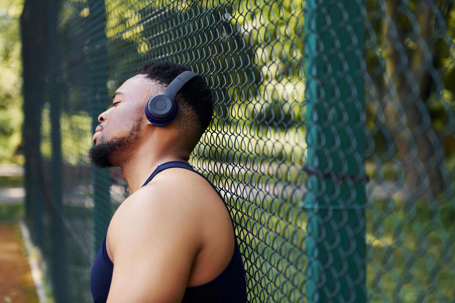 Afroamerikaner mit kabellosen Kopfhörern lehnt sich auf das Metallgitter des Sportplatzes und macht Pause foto