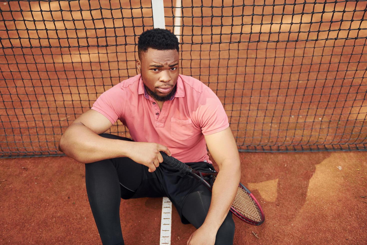 sitzt in der Nähe des Netzes und macht eine Pause. afroamerikanischer mann im rosa hemd sitzt mit tennisschläger auf dem platz im freien foto