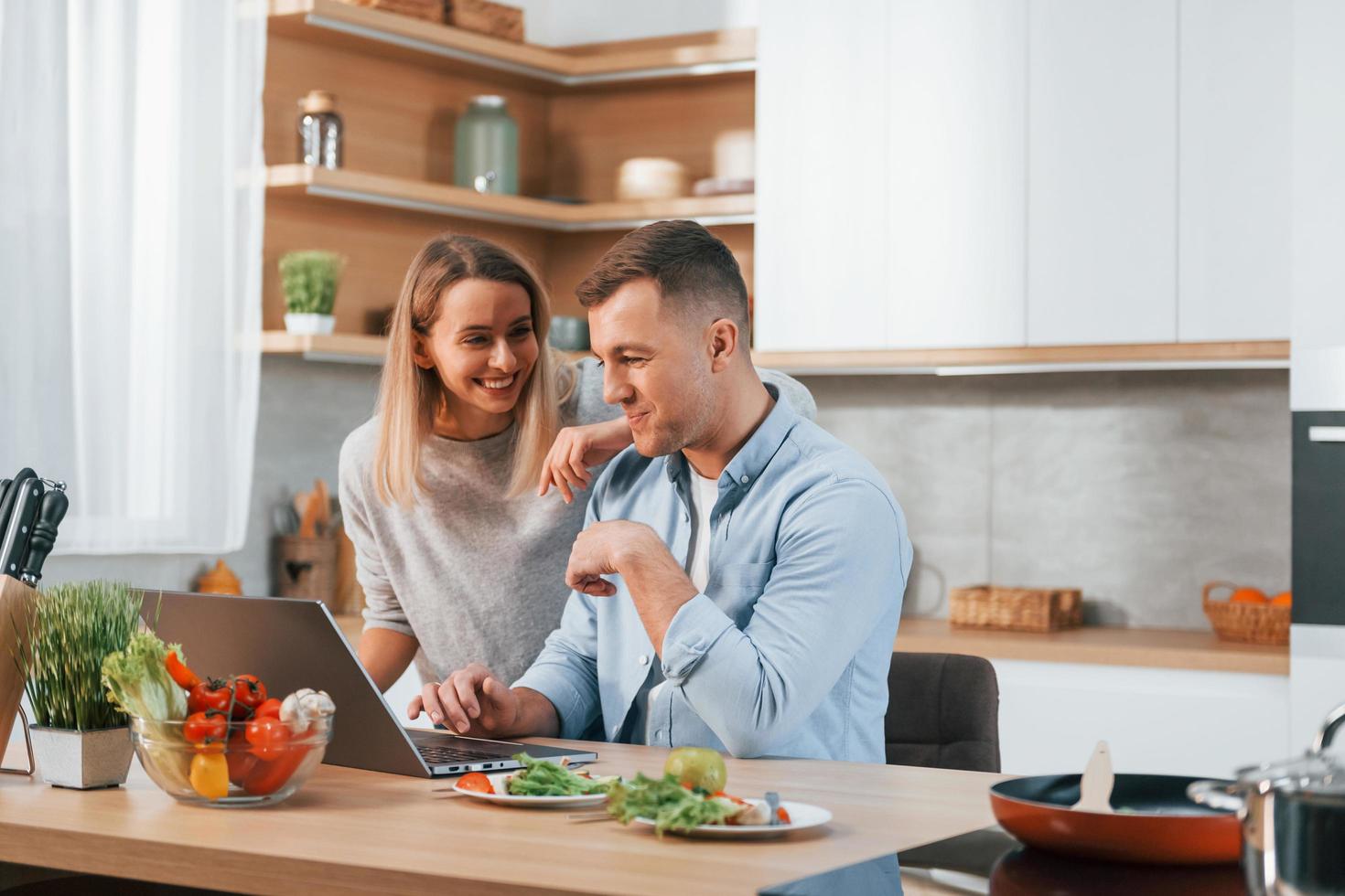 Laptop anschauen. Ehepaar bereitet Essen zu Hause in der modernen Küche zu foto