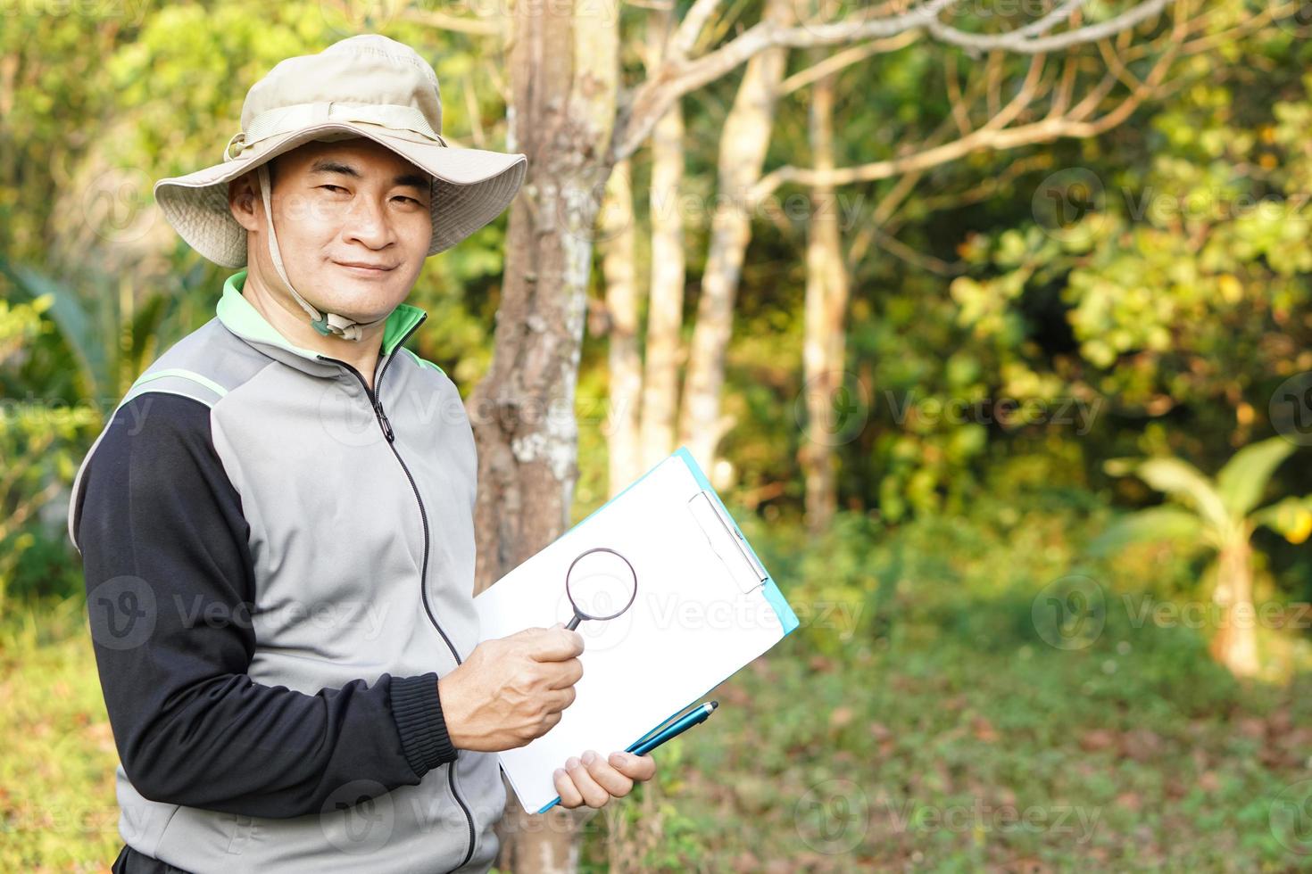 Schöner asiatischer Botaniker ist im Wald, um botanische Pflanzen zu untersuchen, hält Büroklammern und Lupe. über Umwelt, Pflanzen und Natur konzeptionieren, untersuchen, erforschen, forschen, studieren. foto