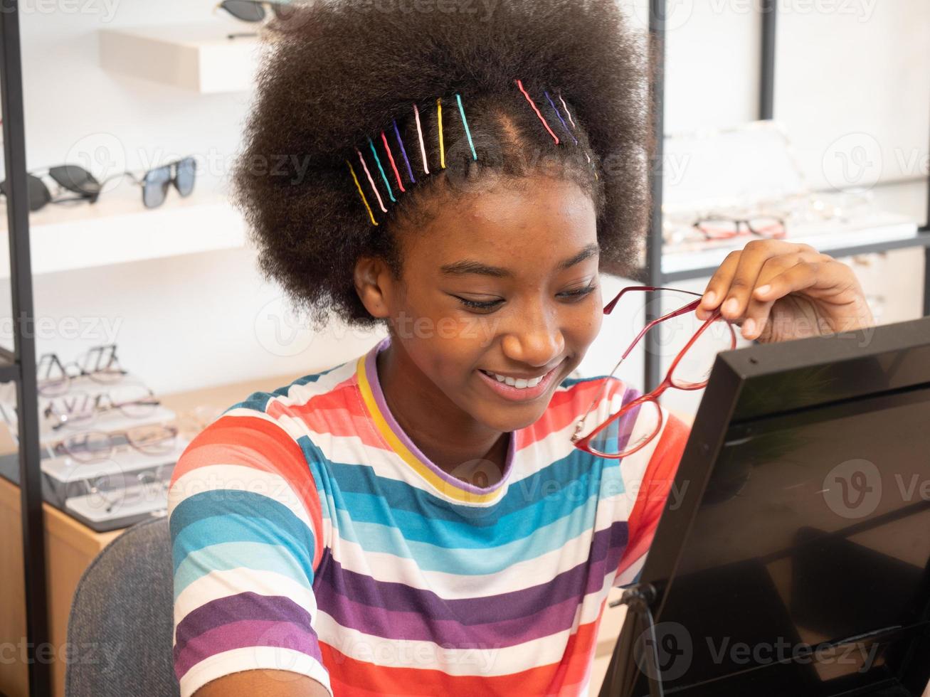 afrikanisches mädchen im optikgeschäft, das in den spiegel schaut, um einen neuen brillenrahmen auszuwählen. Gesundheitsversorgung, Medizin, Menschen, Sehvermögen und Technologiekonzept foto