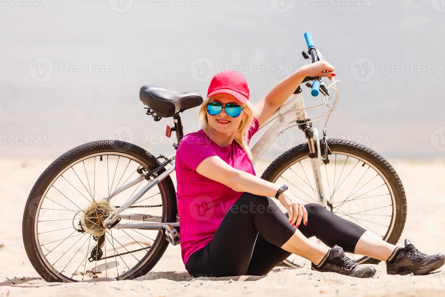 Junges sexy blondes Mädchen mit langen Haaren in Sonnenbrille, das in der Nähe des Fahrrads steht, Spaß hat und gute Laune hat, in die Kamera schaut und lächelt foto