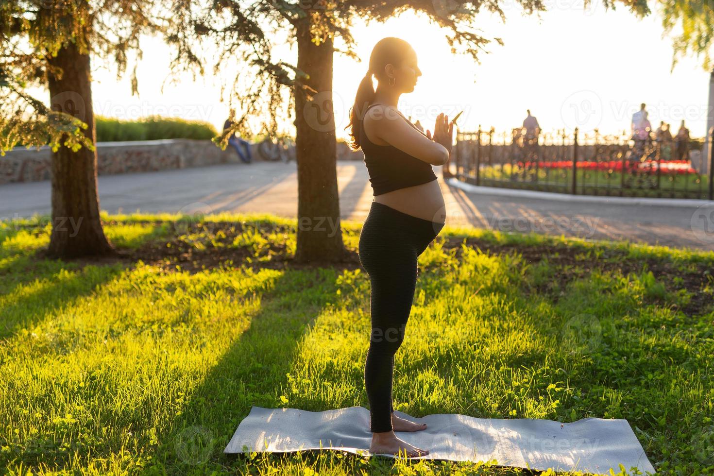 Seitenansicht der jungen Frau, die ein Baby im schwarzen Anzug erwartet, steht barfuß auf dem Teppich und meditiert an einem warmen, sonnigen Sommertag. konzept von sport- und meditationsaktivitäten foto