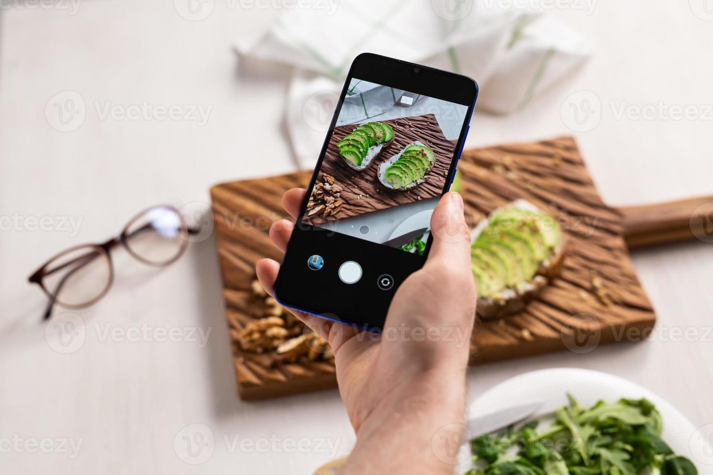 Hände fotografieren auf dem Smartphone zwei schöne, gesunde Sauerrahm- und Avocado-Sandwiches, die an Bord auf dem Tisch liegen. Social Media und Food-Konzept foto