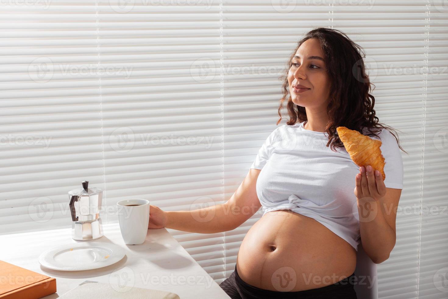 glückliche schwangere junge schöne Frau, die beim morgendlichen Frühstück Croissants isst. konzept eines angenehmen morgens und einer positiven einstellung während der schwangerschaft foto