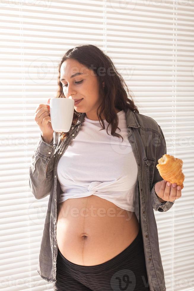 gesunde schöne schwangere frau, die tee trinkt und während des mittagessens croissant isst. konzept der kalorienreichen ernährung während des wartens auf die geburt des babys foto