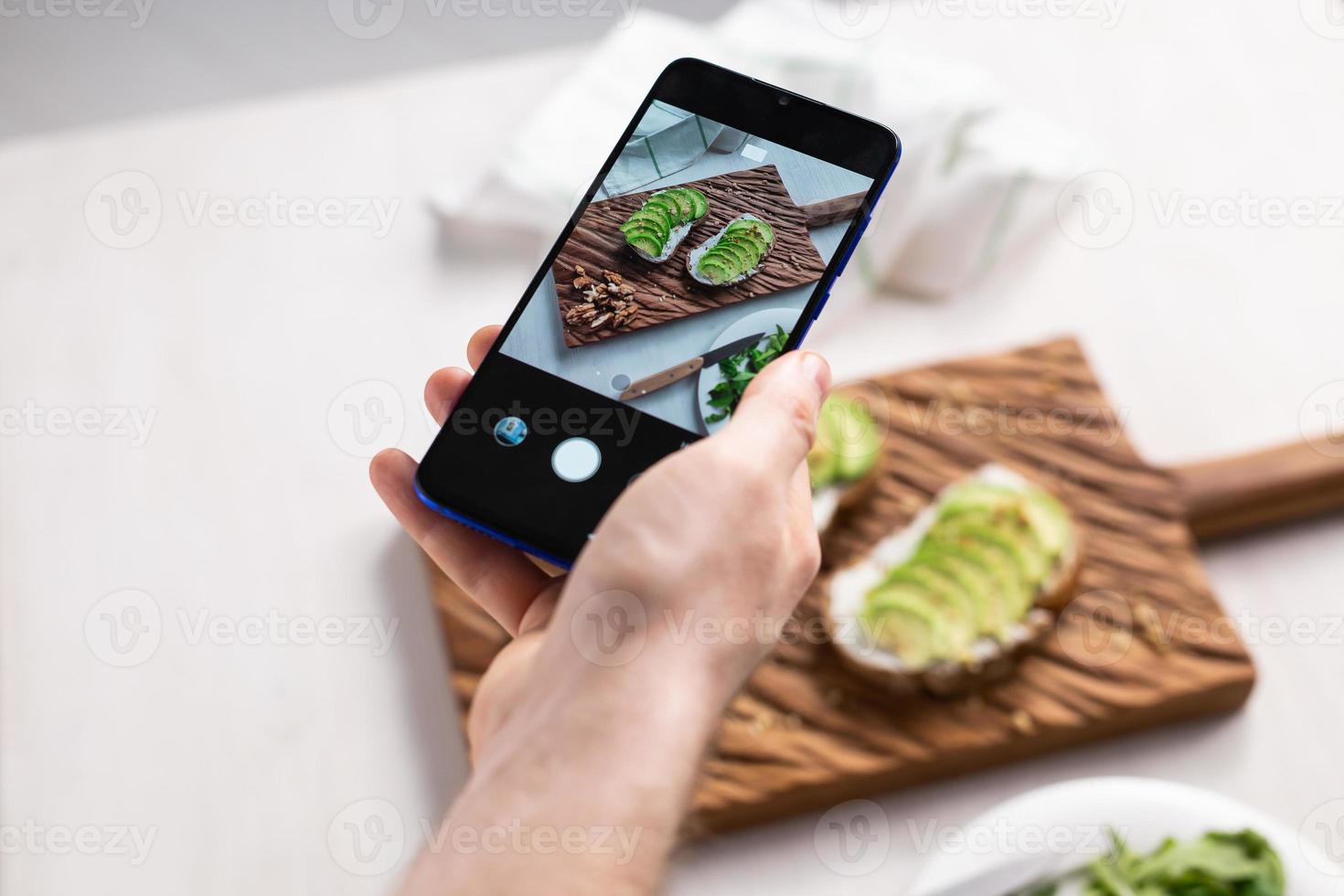 Hände fotografieren auf dem Smartphone zwei schöne, gesunde Sauerrahm- und Avocado-Sandwiches, die an Bord auf dem Tisch liegen. Social Media und Food-Konzept foto