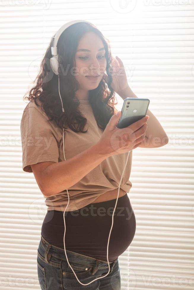 Porträt einer schwangeren Frau in der Nähe des Fensters zu Hause und beim Hören von Musik über Kopfhörer. schwangerschafts- und freizeitkonzept. foto