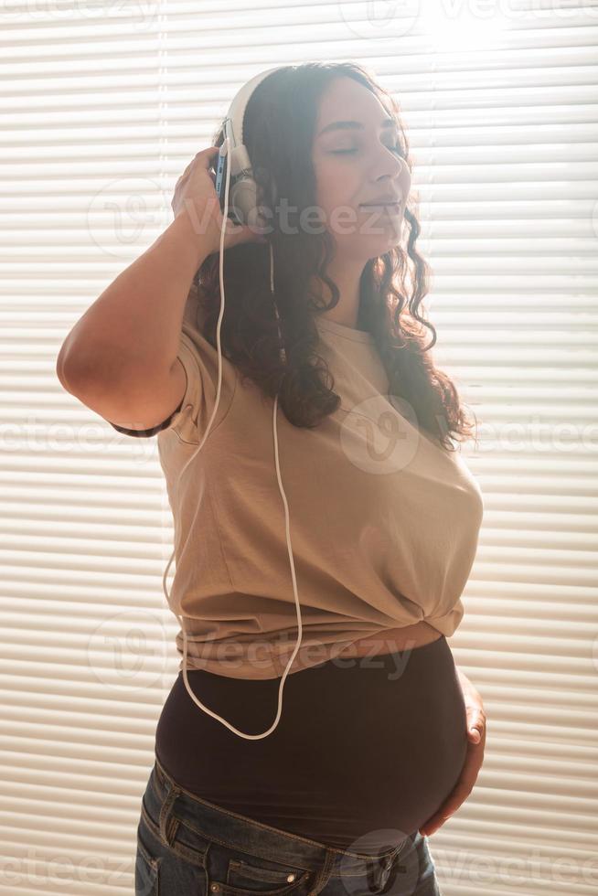 lockige brünette befriedete schwangere frau hört mit smartphone und kopfhörern angenehme klassische musik. konzept einer beruhigenden stimmung vor dem treffen des babys. foto