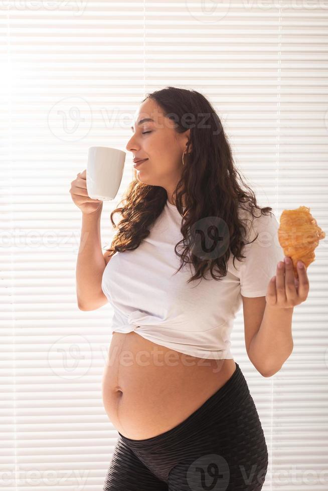 schwangere frau, die croissant isst und kaffee trinkt. Schwangerschaft und Mutterschutz foto