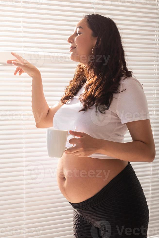 neugierige junge schöne schwangere frau, die tee trinkt und durch die jalousien auf das fenster schaut. konzept der freude und der guten nachrichten beim warten auf das baby foto