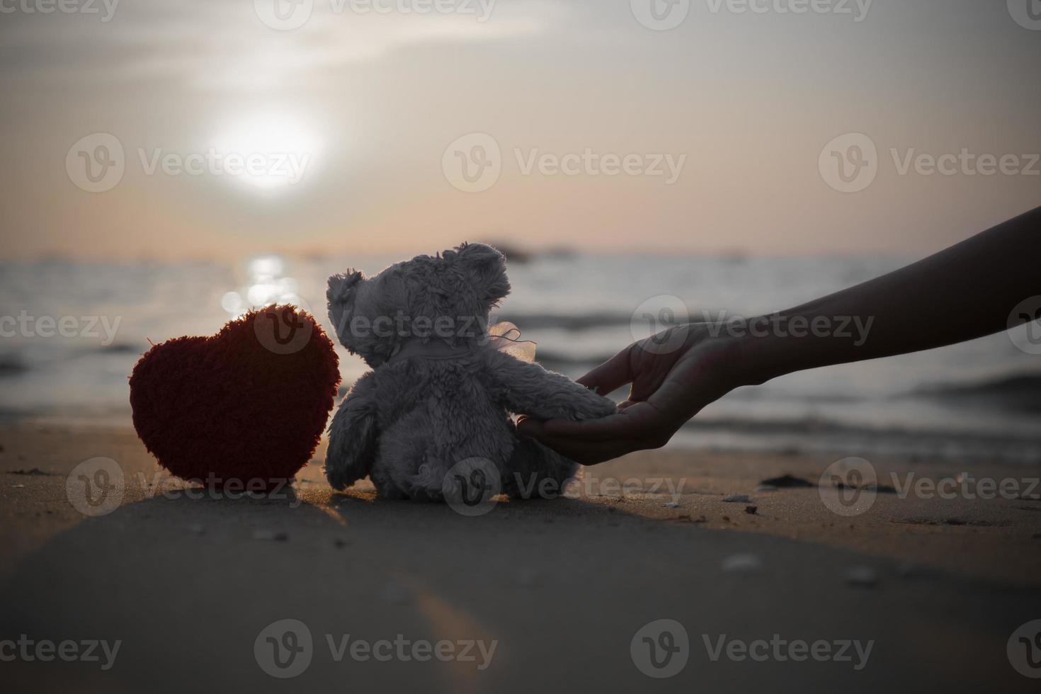 weibliche Hand, die einen kleinen, einsamen, violetten, niedlichen kleinen Bären hält, der mit rotem Handwerksherz am Strand sitzt, um eine Postkarte mit international vermissten Kindern zu erstellen, gebrochenes Herz. foto