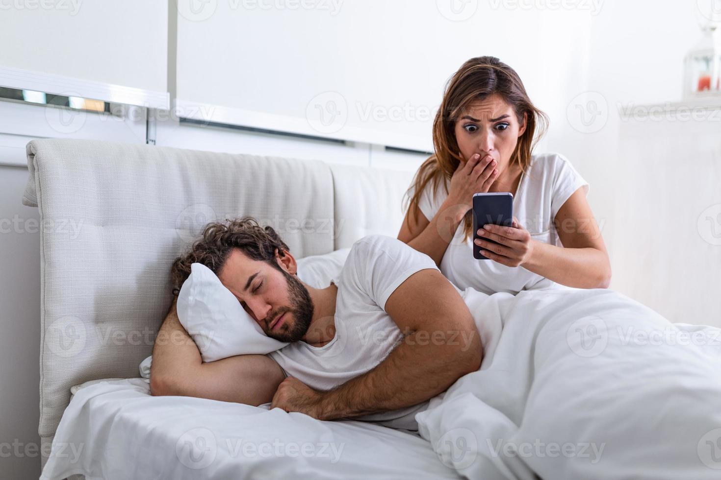 Eifersüchtige Frau, die das Telefon ihres Partners ausspioniert, während er zu Hause in einem Bett schläft. Schockierte eifersüchtige Frau, die das Telefon ihres Mannes ausspioniert, während der Mann zu Hause im Bett schläft foto