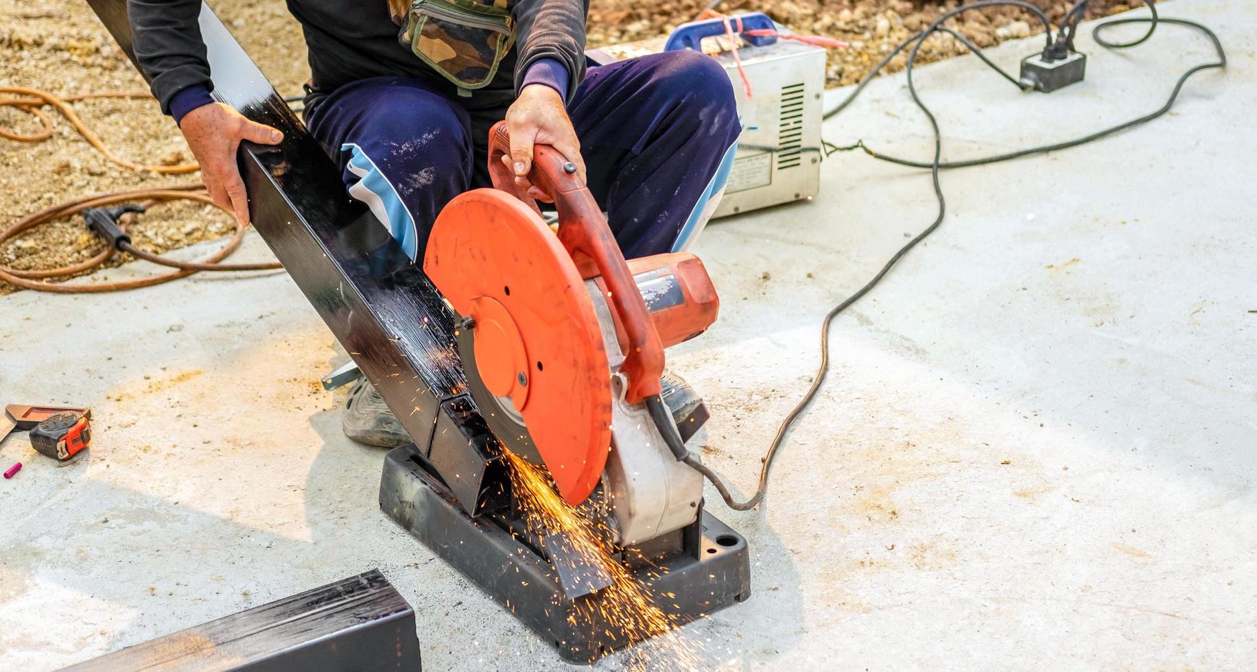 Der Arbeiter in der Arbeitskleidung verwendet einen elektrischen Stahlschneider. Schneiden von großen Stahlstangen auf der Baustelle foto