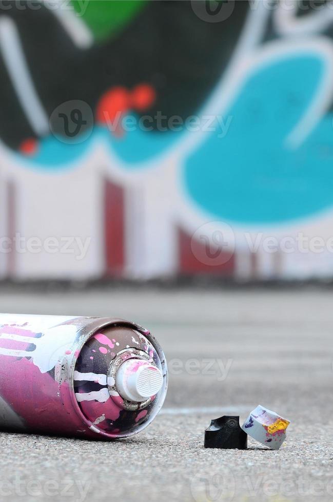 Gebrauchte Aerosol-Sprühdose mit rosa und weißer Farbe und Kappen zum Sprühen von Farbe unter Druck liegt auf dem schmutzigen Asphalt in der Nähe der bemalten Wand in farbigen Graffiti-Zeichnungen foto