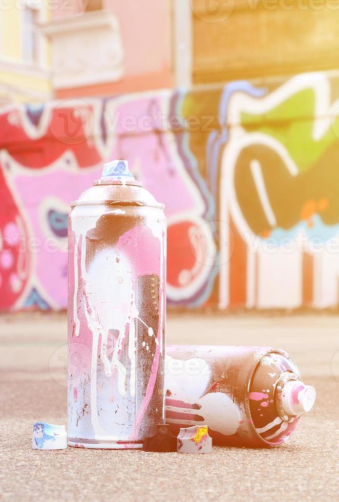 mehrere gebrauchte sprühdosen mit rosa und weißer farbe und kappen zum sprühen von farbe unter druck liegen auf dem asphalt in der nähe der bemalten wand in farbigen graffiti-zeichnungen foto