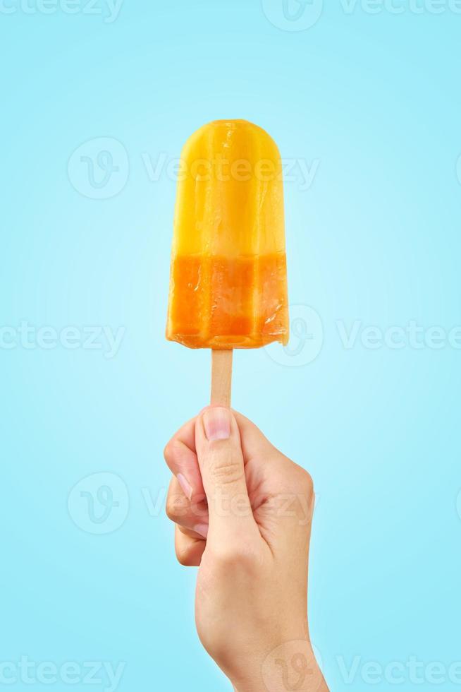 gelbes gefrorenes fruchteis am stiel in der hand auf blauem hintergrund foto