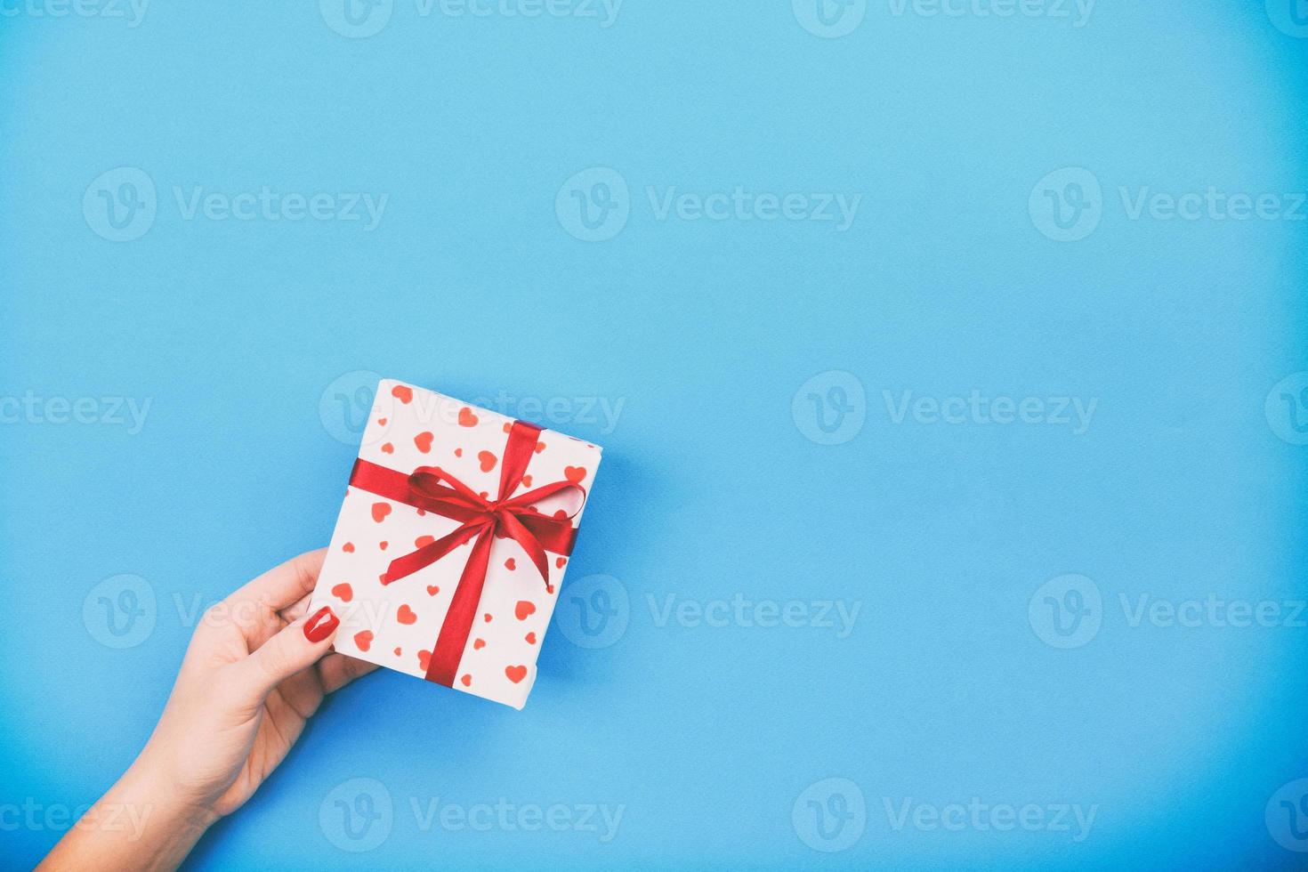 Frauenhände geben eingewickeltes Valentinsgruß- oder anderes Feiertagshandgemachtes Geschenk im Papier mit rotem Band. geschenkbox, rote herzdekoration des geschenks auf blauem tisch, draufsicht mit kopierraum für ihr design foto
