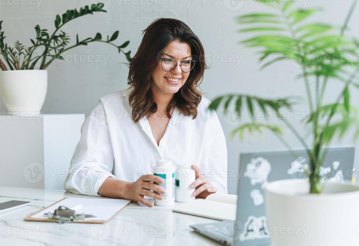 Junge lächelnde brünette Frau Ernährungsberaterin plus Größe in weißem Hemd, die am Laptop auf dem Tisch mit Zimmerpflanze im hellen, modernen Büro arbeitet. Der Arzt kommuniziert online mit dem Patienten foto