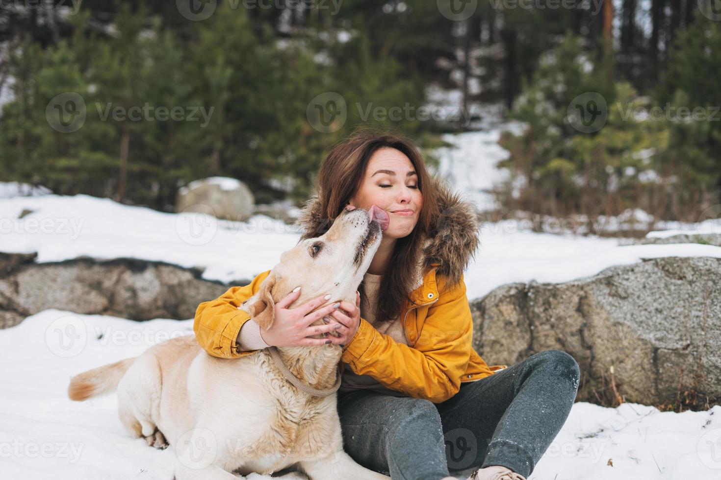 Junge lächelnde Frau in gelber Jacke mit großem freundlichen weißen Hund Labrador, der im Winterwald spazieren geht foto