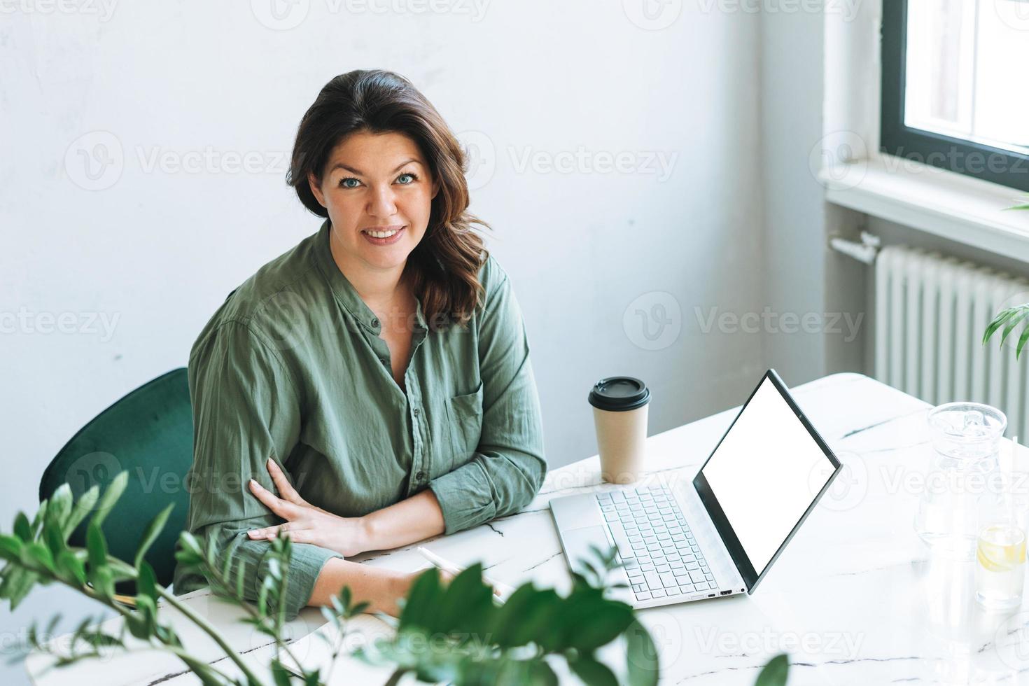 junge lächelnde brünette frau plus größe, die am laptop mit weißem bildschirm auf dem tisch mit zimmerpflanze im hellen modernen büro arbeitet foto