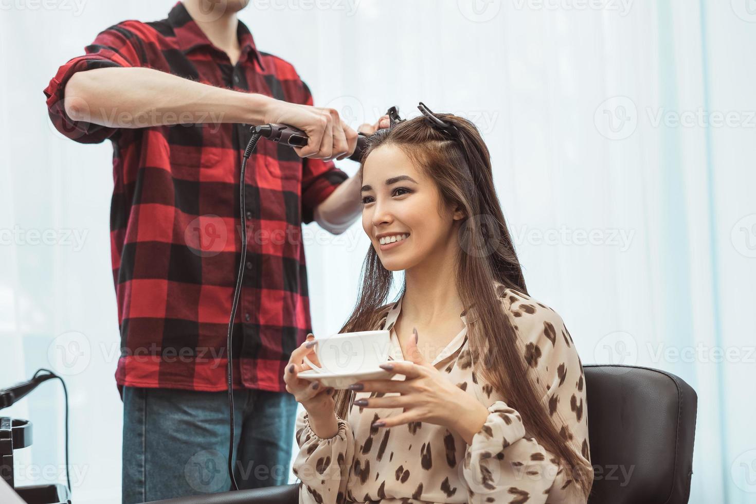 friseurfriseur, der lange haare mit haareisen für schöne junge frau im schönheitssalon stylt, arbeitsmoment foto