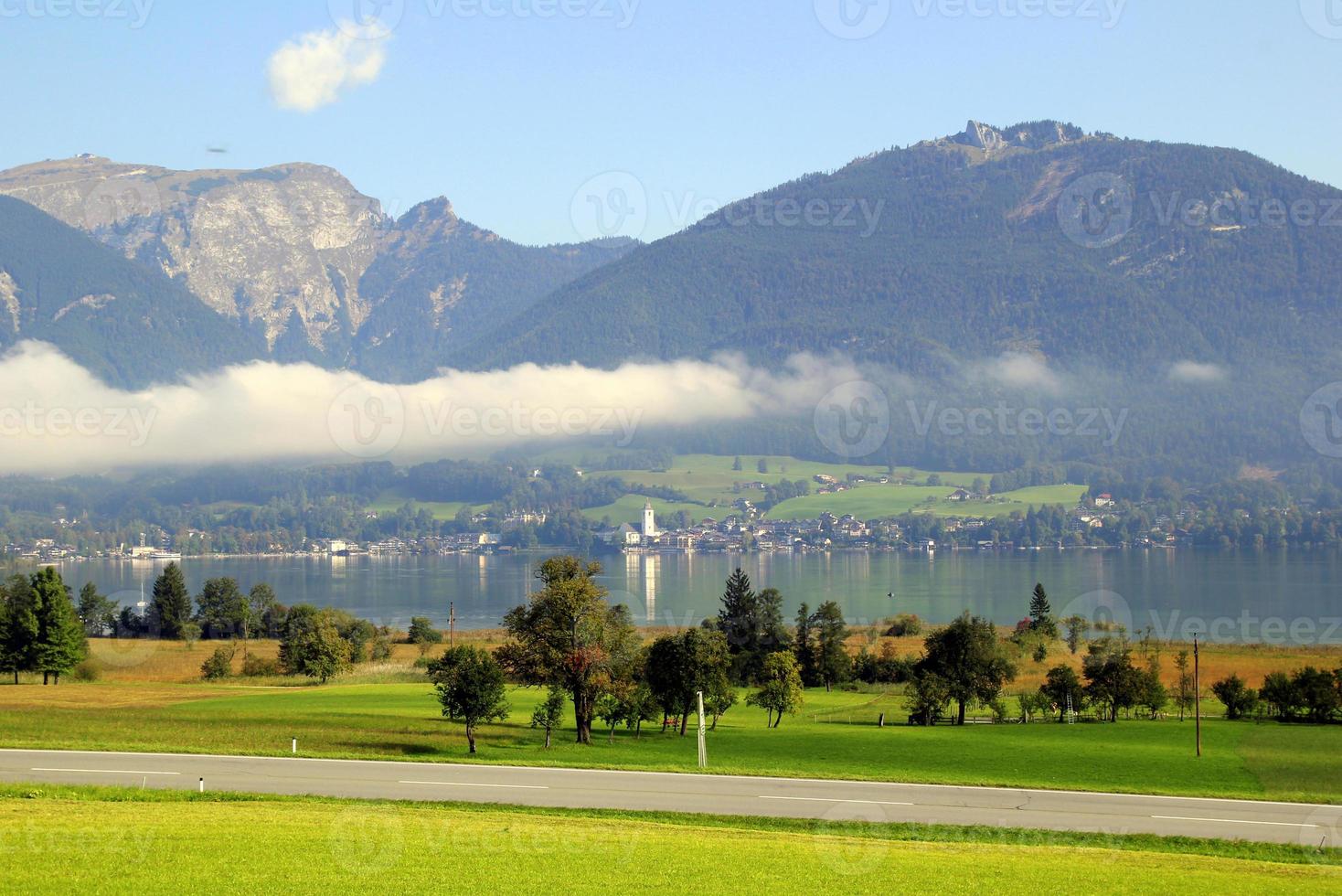 reise nach sankt-wolfgang, österreich. die straße zwischen grünen wiesen mit den bergen und einem see in den wolken im hintergrund am sonnigen tag. foto