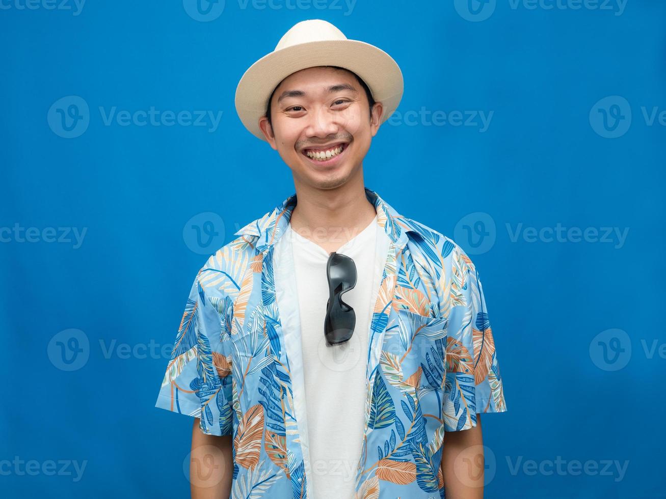 reisender asiatischer mann trägt hut mit sonnenbrille glücklichem lächeln blauem hintergrund foto