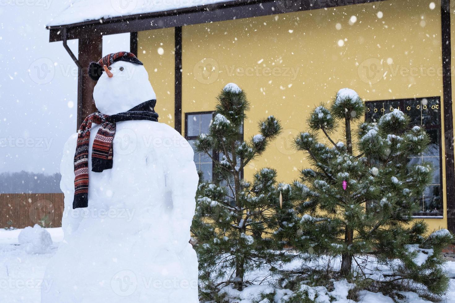 Lustiger Schneemann mit Hut und Schal auf dem Hintergrund eines gelben Hauses im Hof. Winter, Winterunterhaltung, Schneefall foto
