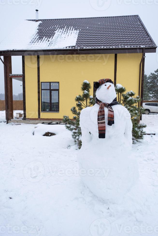 Lustiger Schneemann mit Hut und Schal auf dem Hintergrund eines gelben Hauses im Hof. Winter, Winterunterhaltung, Schneefall foto