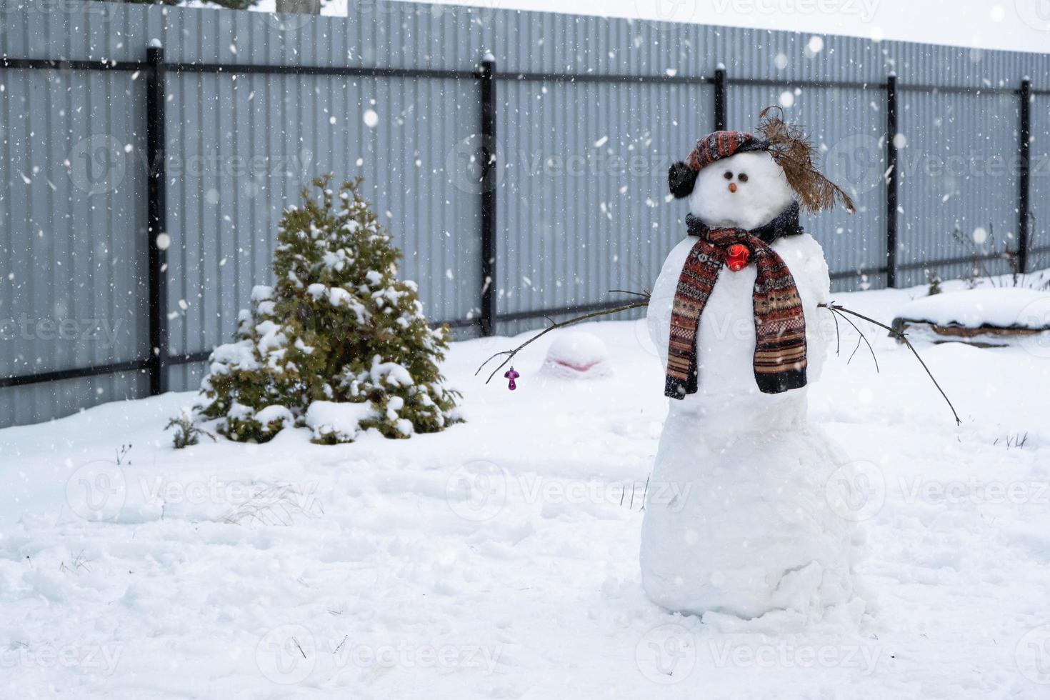 Lustiger Schneemann mit Hut und Schal auf dem Hintergrund eines gelben Hauses im Hof. Winter, Winterunterhaltung, Schneefall foto