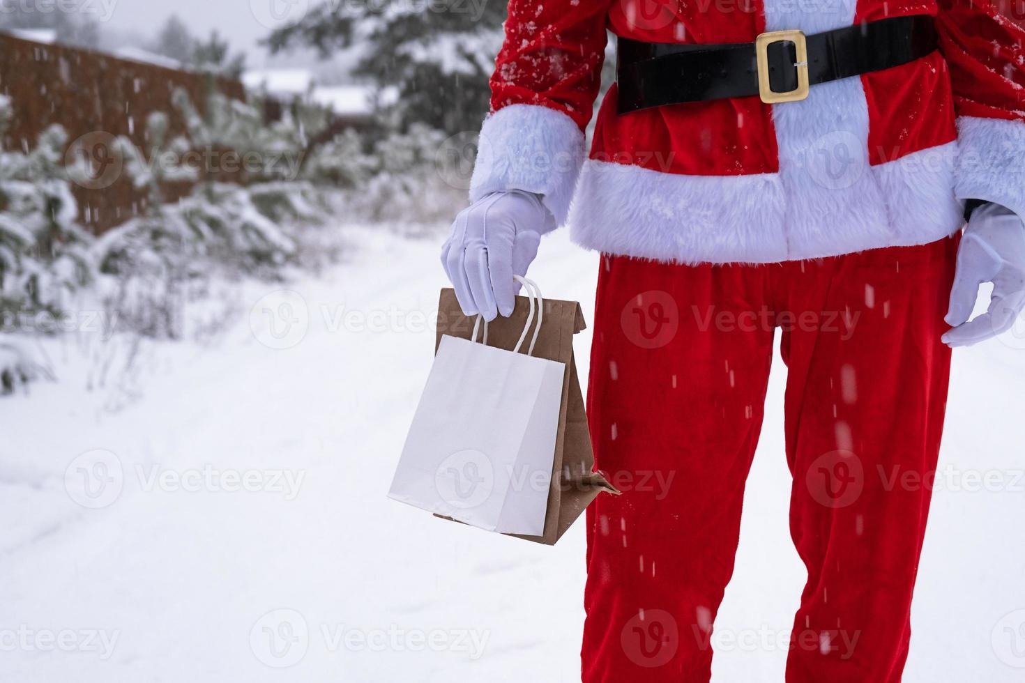 weihnachtsmann im freien im winter und schnee, der handpapiertüten mit handwerksgeschenk, lebensmittellieferung übergibt. Einkaufen, Verpackungsrecycling, Handarbeit, Lieferung für Weihnachten und Neujahr foto