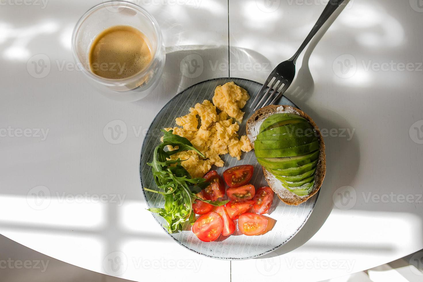 Teller mit Rührei, Rucola, Tomate, Vollkorntoast mit Avocado, Tasse Kaffee auf dem Tisch. foto