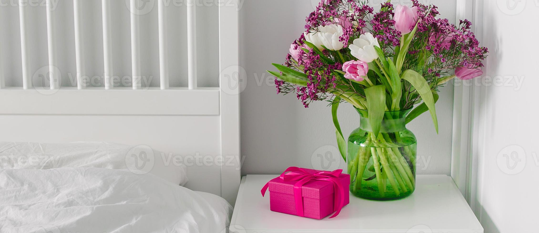 Rosa Geschenkbox und Vase mit Frühlingsblumen auf dem Nachttisch neben dem Bett. skandinavisches Interieur. foto
