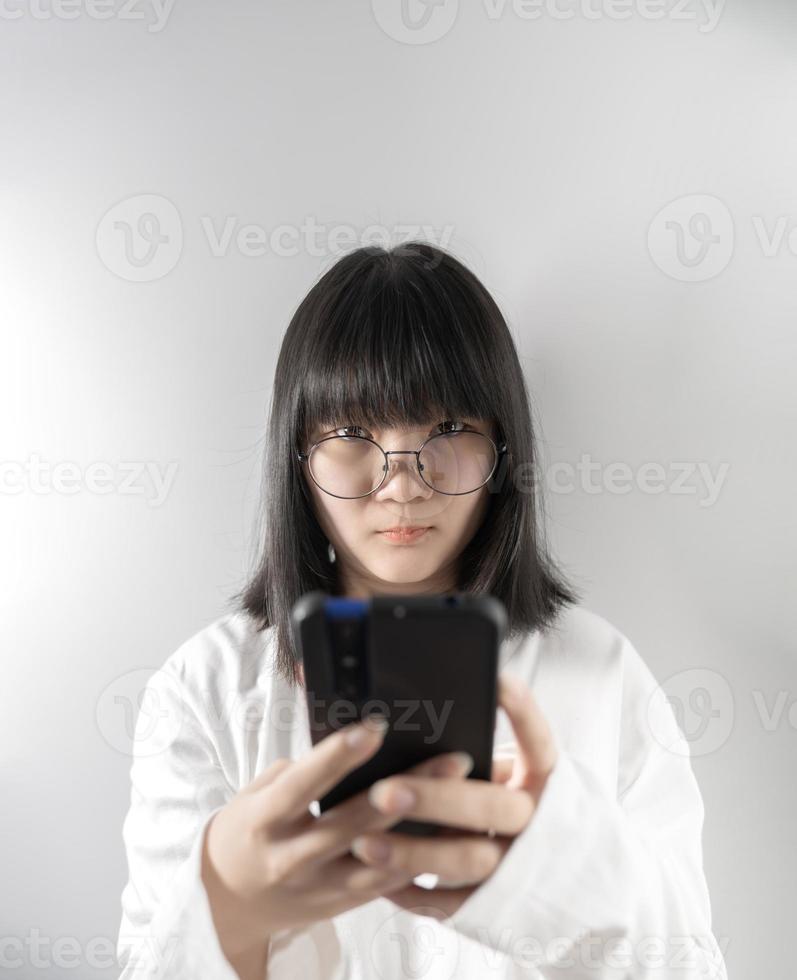 isoliert hübsche asiatische brille frau schaut in die kamera und hält handy in der hand auf weißem hintergrund im studiolicht. foto